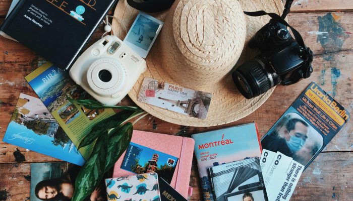 Flat lay of travel accessories including a straw hat, cameras, and guidebooks on a wooden table.